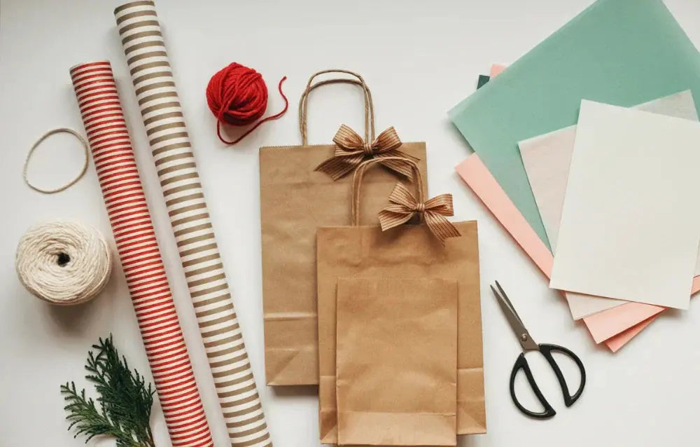 Gift wrapping supplies arranged on a white table including kraft bags, ribbon, wrapping paper rolls, scissors, twine and colored paper.