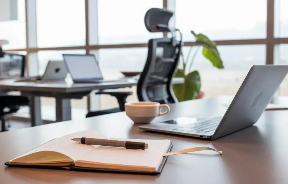 Height-adjustable standing desk positioned by a window with monitor, keyboard, notebook, and ceramic mug, bright natural light.